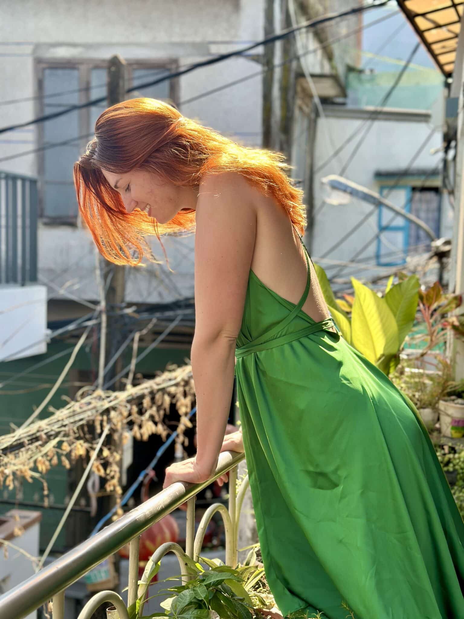 Woman in a green dress standing on a balcony with buildings and plants in the background