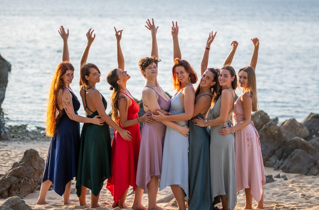 Group of women posing on a beach with arms raised, wearing colorful dresses.