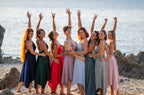 Group of women posing on a beach with arms raised, wearing colorful dresses.